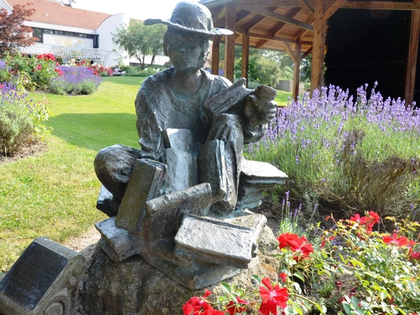 Lesende_HD_16zu9.jpg Bronzestatue Die Lesende auf Büchern in einem Garten mit Lavendel und roten Rosen.