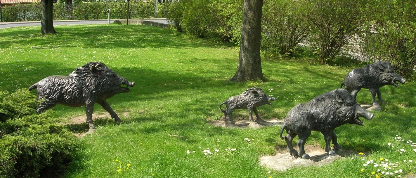 Wildschweine_HD_16zu9.JPG Vier lebensechte Bronze-Wildschweine stehen in dynamischen Posen auf einer grünen Rasenfläche.