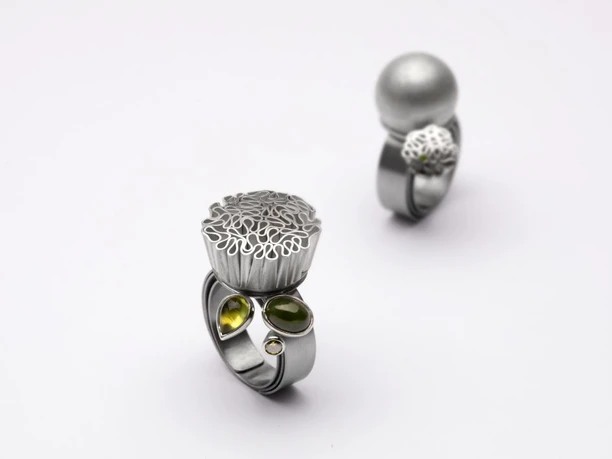 Collaboration between Jutta Grote and Sabine Braunfisch Two silver designer rings with filigree attachments and green gemstones against a light background.