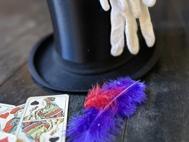 Zauberwerkstatt Zylinder mit weißen Handschuhen, Spielkarten und bunten Federn auf dunklem Holz.