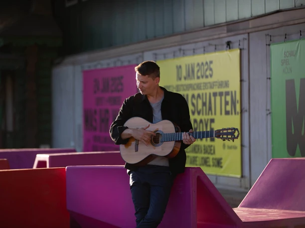 Daniel Stelter-2-(c) Victoria-Page.jpg Daniel Stelter spielt Gitarre vor großer bunter Plakatwand