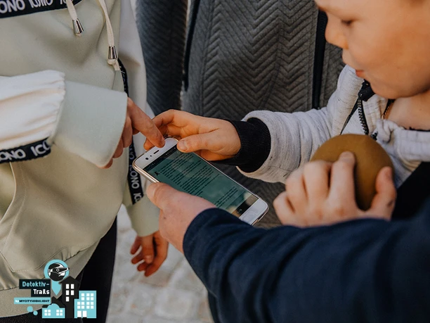 Detektiv-Trails-Familie-Schatzsuche-Spiel (c) MyCityHighlight Europe GmbH.jpg Ein Kind zeigt auf ein Smartphone in den Händen eines Erwachsenen bei einer Schatzsuche.
