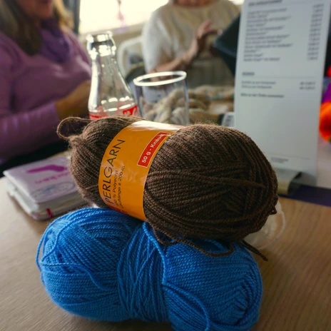 Stikke-Kaffee - Woll-Kneule.JPG Zwei Wollknäuel in Blau und Braun auf einem Tisch. Im Hintergrund sitzen zwei Frauen.