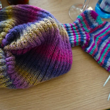 Strikke-Kaffee - Mütze.JPG Bunte gestrickte Mütze und ein halbfertiger Strumpf mit Stricknadeln auf einem Holztisch.