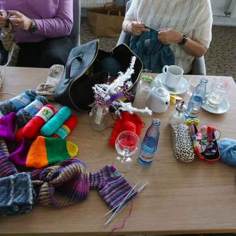 Strikke-Kaffee - Titelbild.JPG Menschen stricken an einem Tisch voller bunter Wolle, Strickzeug und Getränken.