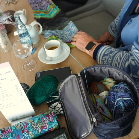 Stikke-Kaffee - Drachen-Schal.JPG Frauen stricken gemeinsam am Tisch mit Tassen, bunter Wolle und einem Strickschal.