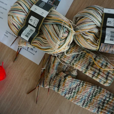 Stikke-Kaffee - Woll u Socken.JPG Bunte Wollknäuel, Stricknadeln und halbfertige Socken liegen auf einem Holztisch.