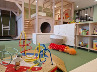 In the foreground you can see a play table with a motor skills loop, in the background a cube shelf with books and other toys and a two-storey indoor playground with stairs and swings