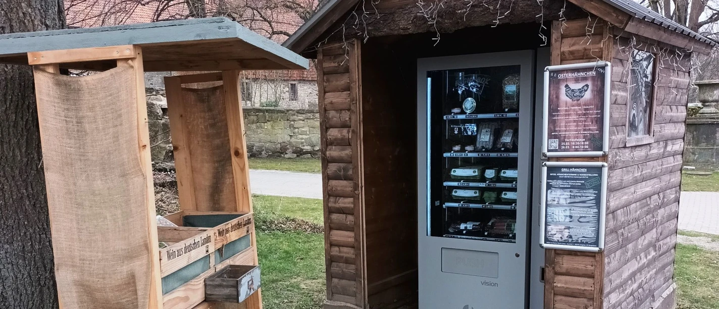 Regiomat_Gutshof_Löwe_HD_16zu9.jpg Regiomat in kleiner Holzhütte neben einem Holzstand mit regionalen Produkten im Freien.