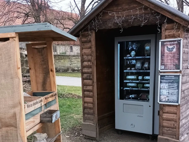 Regiomat_Gutshof_Löwe_HD_16zu9.jpg Regiomat in kleiner Holzhütte neben einem Holzstand mit regionalen Produkten im Freien.