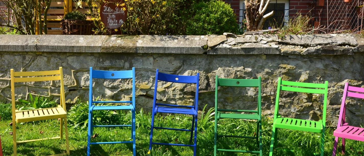 Fruchtaufstrich_Ittermann_HD_16zu9.JPG Sechs bunte Klappstühle stehen auf einer grünen Wiese direkt vor einer Steinmauer.