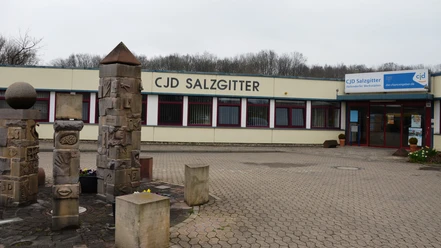 CJD Hallendorf_HD_16zu9.JPG Flaches Gebäude des CJD Salzgitter mit gepflastertem Vorplatz und verzierten Steinsäulen.