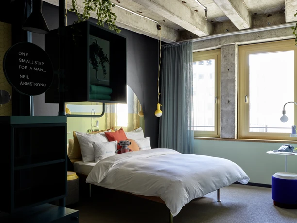 25hours Hotel The Circle Zimmer Modernes Hotelzimmer mit Doppelbett, großen Fenstern, Pflanzen und farbigen Designelementen.