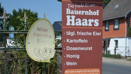 Hofladen_Haars_HD_16zu9.JPG Ein braunes Schild vor einem Hoftor bewirbt frische Eier, Kartoffeln und Honig vom Bauernhof Haars.