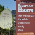 Hofladen_Haars_HD_16zu9.JPG Ein braunes Schild vor einem Hoftor bewirbt frische Eier, Kartoffeln und Honig vom Bauernhof Haars.