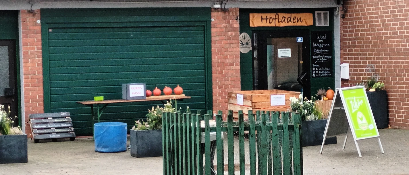 Hofladen_Hagemann_Engelnstedt_HD_16zu9.jpg Backstein-Hofladen mit Tür, Kürbissen auf einem Tisch und grünem Zaun im Vordergrund.