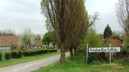Zufahrt zu Fachwerkhäusern. Ein Schild am Wegrand bewirbt frische Kartoffeln.
