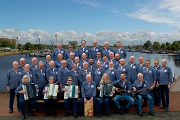 Fehntjer-Shanty-Chor.jpg Fehntjer Shanty Chor in blauer Kleidung mit Instrumenten vor einem Hafen mit Booten.