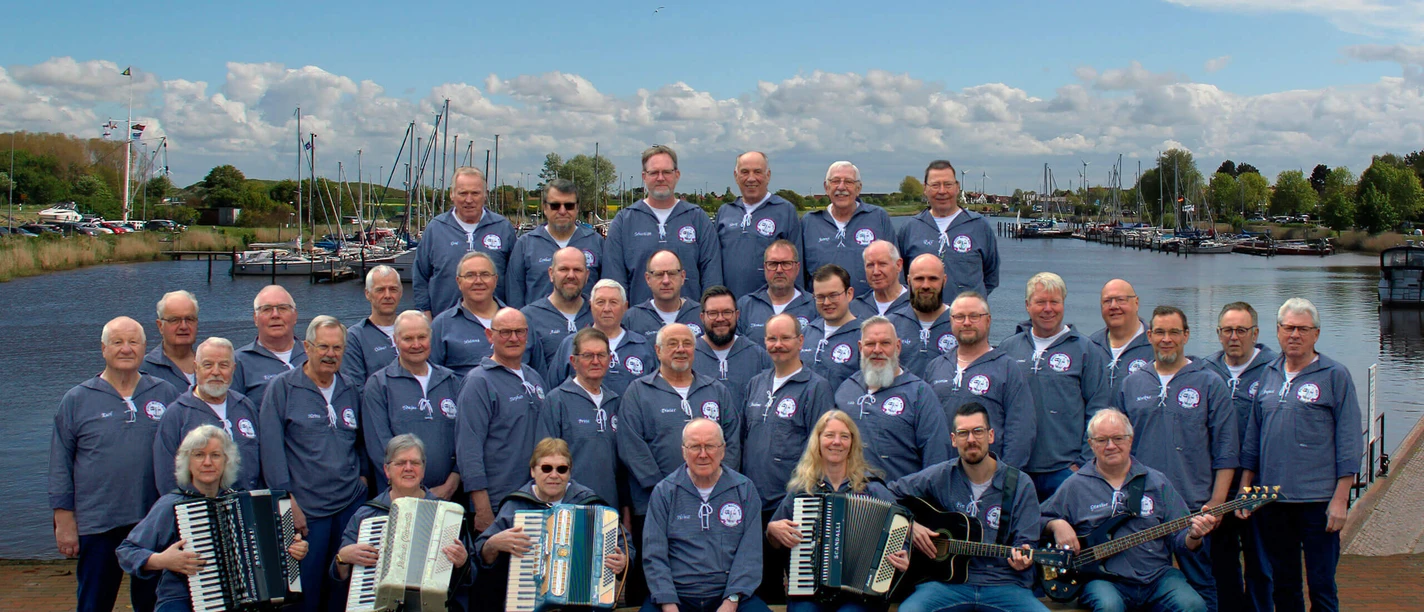 Fehntjer-Shanty-Chor.jpg Fehntjer Shanty Chor in blauer Kleidung mit Instrumenten vor einem Hafen mit Booten.
