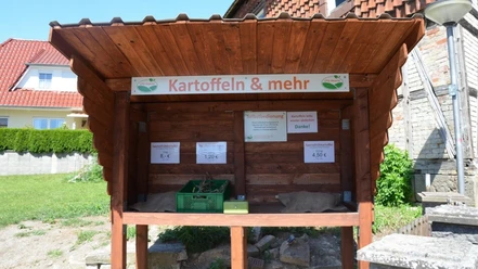 Ein hölzerner Verkaufsstand zur Selbstbedienung mit Schild Kartoffeln & mehr.