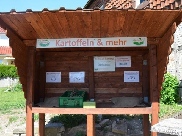 Ein hölzerner Verkaufsstand zur Selbstbedienung mit Schild Kartoffeln & mehr.