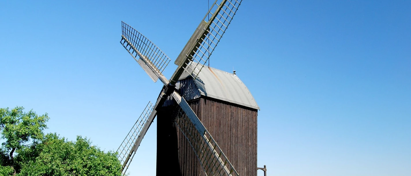 Mühle Lichtenberg_Anne König_HD_16zu9.jpg Historische Bockwindmühle aus Holz mit Flügeln vor blauem Himmel und grünen Bäumen.