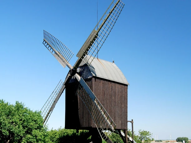 Mühle Lichtenberg_Anne König_HD_16zu9.jpg Historische Bockwindmühle aus Holz mit Flügeln vor blauem Himmel und grünen Bäumen.