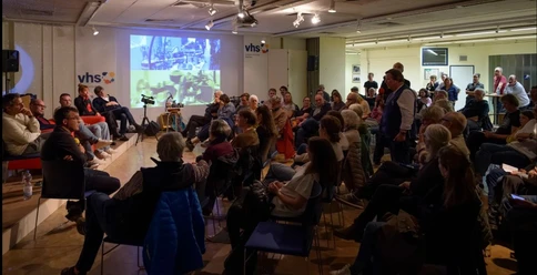 In einem kleinen Saal wird ein Vortrag mit Präsentation vor einem sitzenden Publikum gehalten.