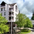 Außenansicht, Apartment - Der Gerber in Hilden Moderne Außenansicht des Apartment - Der Gerber in Hilden. Mehrere Bäume säumen den Gehweg, ein Kirchturm ragt im Hintergrund auf.