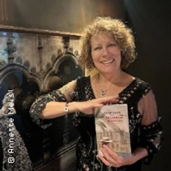 Eine Frau mit lockigem Haar hält ein Buch und lächelt vor einer dunklen Wand.