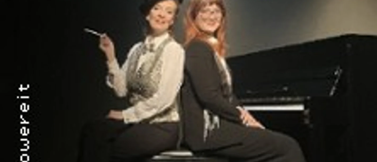 Zwei Personen sitzen Rücken an Rücken vor einem Klavier im Scheinwerferlicht.