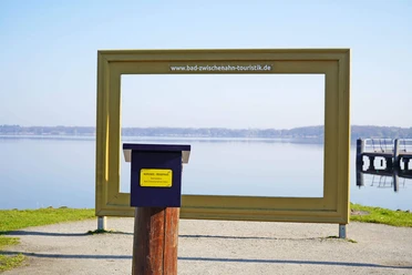 Stempelstelle-TANO-Reisepass-Goldener-Bilderrahmen-Bad -Zwischenahn.jpg Ein großer goldener Bilderrahmen steht am Ufer des Zwischenahner Meeres, davor ein Stempelkasten.