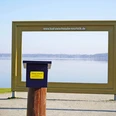 Stempelstelle-TANO-Reisepass-Goldener-Bilderrahmen-Bad -Zwischenahn.jpg Ein großer goldener Bilderrahmen steht am Ufer des Zwischenahner Meeres, davor ein Stempelkasten.