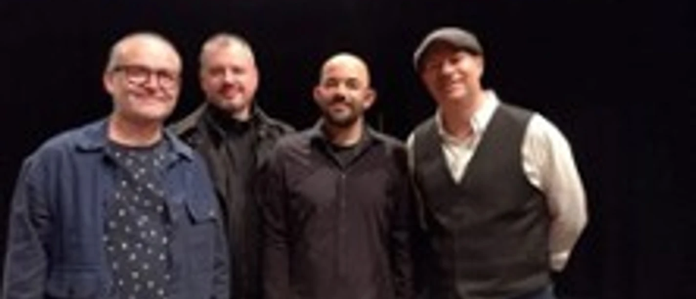 Vier Musiker stehen nebeneinander vor dunklem Hintergrund und lächeln in die Kamera.