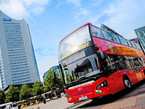 A red double-decker bus from the Original Leipzig City Tours is parked on Augustusplatz, with the City Tower in the background
