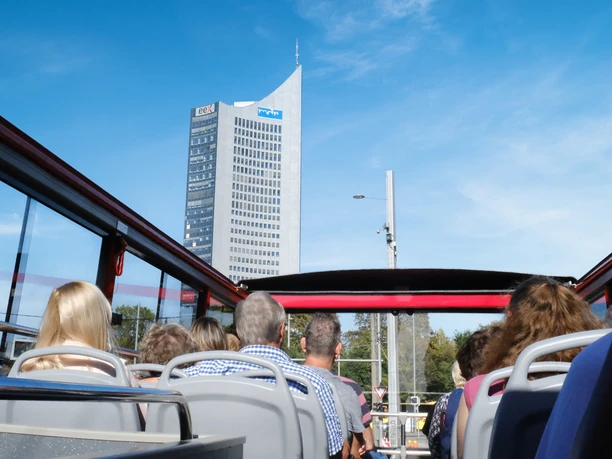 Widok z tylnych rzędów górnego pokładu piętrowego autobusu Original Leipzig City Tours na osoby siedzące z przodu i widok na wieżowiec na Augustusplatz.