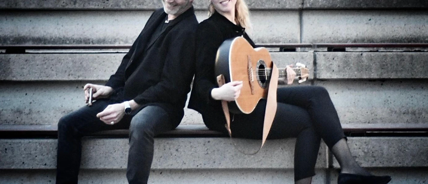 Kultur.Sommer.Garten. mit Anne & Frank Ein Mann und eine Frau sitzen auf einer Tribüne. Die Frau hält eine Gitarre in der Hand.