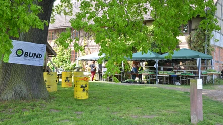 BUND_Salzgitter_Waldhaus_Altenhagen_HD_16zu9.JPG BUND-Banner an großem Baum, gelbe Protestfässer und Pflanzenstände vor einem Fachwerkhaus.