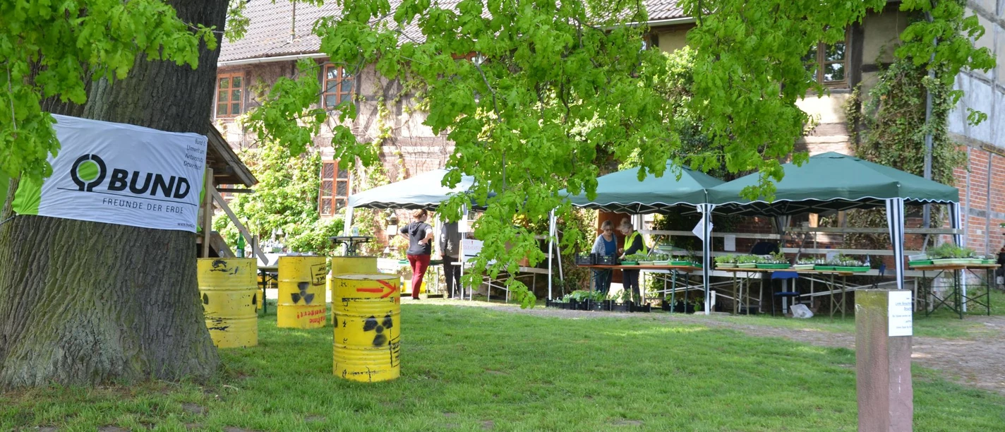 BUND_Salzgitter_Waldhaus_Altenhagen_HD_16zu9.JPG BUND-Banner an großem Baum, gelbe Protestfässer und Pflanzenstände vor einem Fachwerkhaus.