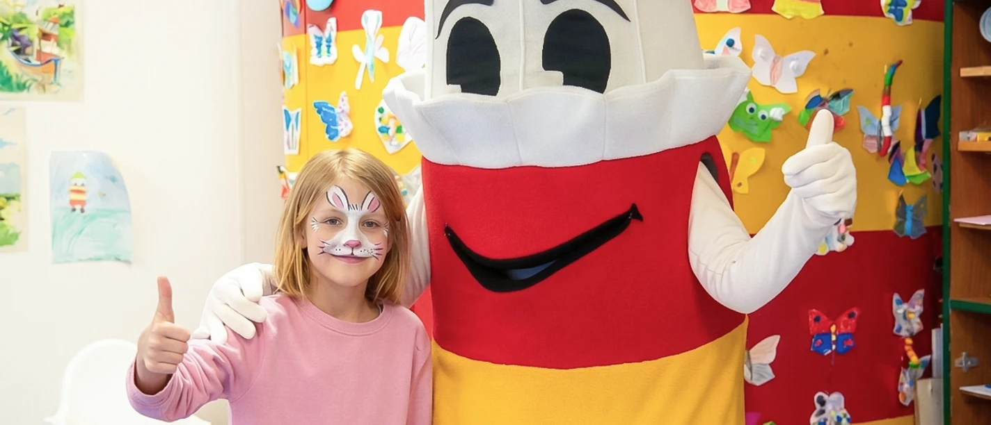Maskottchen Lükko Leuchtturm im Kinderhaus Ein lachendes Kind mit Hasen-Gesichtsbemalung steht neben dem Leuchtturm-Maskottchen Lükko.