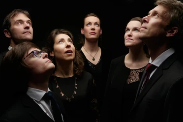 Kölner Vokalsolisten Six formally dressed adults stand in low light, gazing upward with focused, calm expressions.