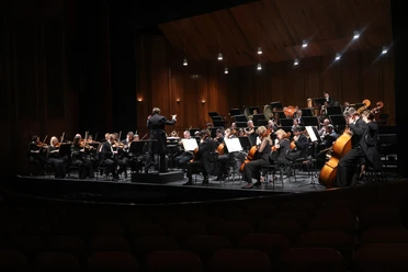 PHILHARMONISCHES ORCHESTER ON ICE_WEB.jpg Ein großes Orchester spielt auf einer beleuchteten Holzbühne, davor leere Zuschauerreihen.