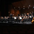 PHILHARMONISCHES ORCHESTER ON ICE_WEB.jpg Ein großes Orchester spielt auf einer beleuchteten Holzbühne, davor leere Zuschauerreihen.