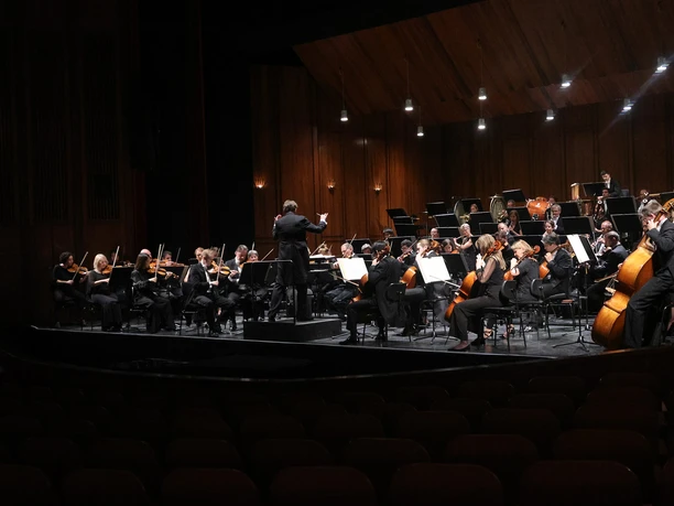 PHILHARMONISCHES ORCHESTER ON ICE_WEB.jpg Ein großes Orchester spielt auf einer beleuchteten Holzbühne, davor leere Zuschauerreihen.