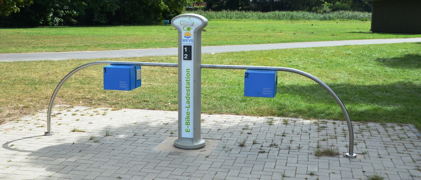 E-Bike-Ladestelle_Salzgittersee_WEVG_HD_16zu9.JPG Moderne E-Bike-Ladestation am Salzgittersee mit vier Anschlüssen auf gepflasterter Fläche im Grünen.