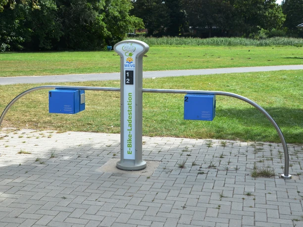 E-Bike-Ladestelle_Salzgittersee_WEVG_HD_16zu9.JPG Moderne E-Bike-Ladestation am Salzgittersee mit vier Anschlüssen auf gepflasterter Fläche im Grünen.