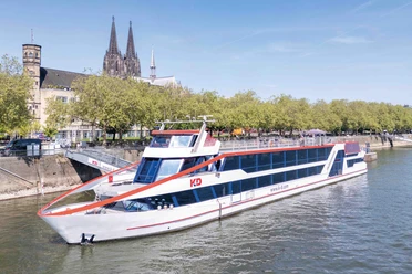 MS RheinDiamond Modern KD Rhine ship in front of the Cologne waterfront with trees and the cathedral in the background
