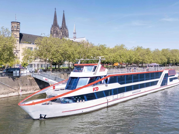 MS RheinDiamond Modern KD Rhine ship in front of the Cologne waterfront with trees and the cathedral in the background