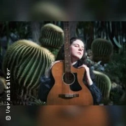 Person hält eine Akustikgitarre vor sich, umgeben von großen Kakteen im Hintergrund