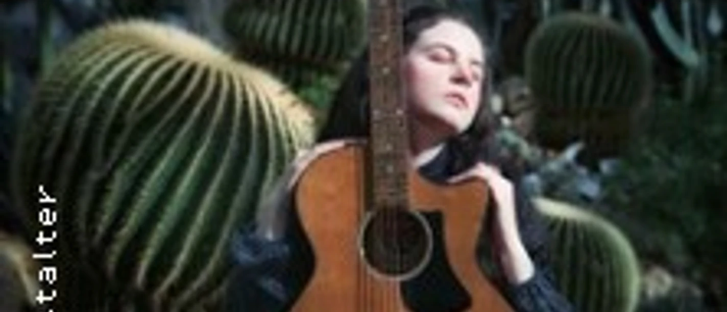 Person hält eine Akustikgitarre vor sich, umgeben von großen Kakteen im Hintergrund